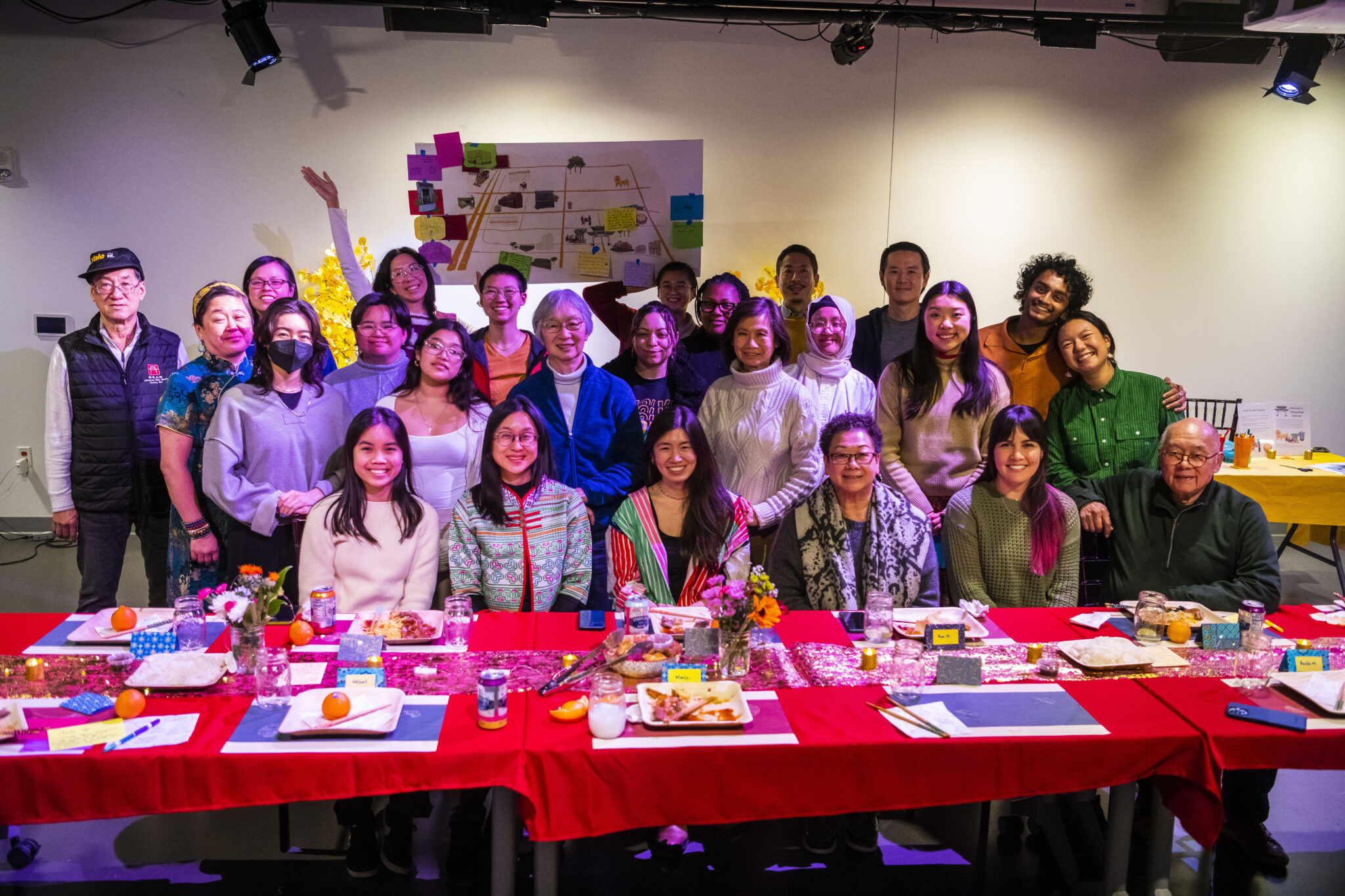 Photo of participants in Memory Mapping Dinner, posing in front of a red tablecloth and holding a map with their memories on it.